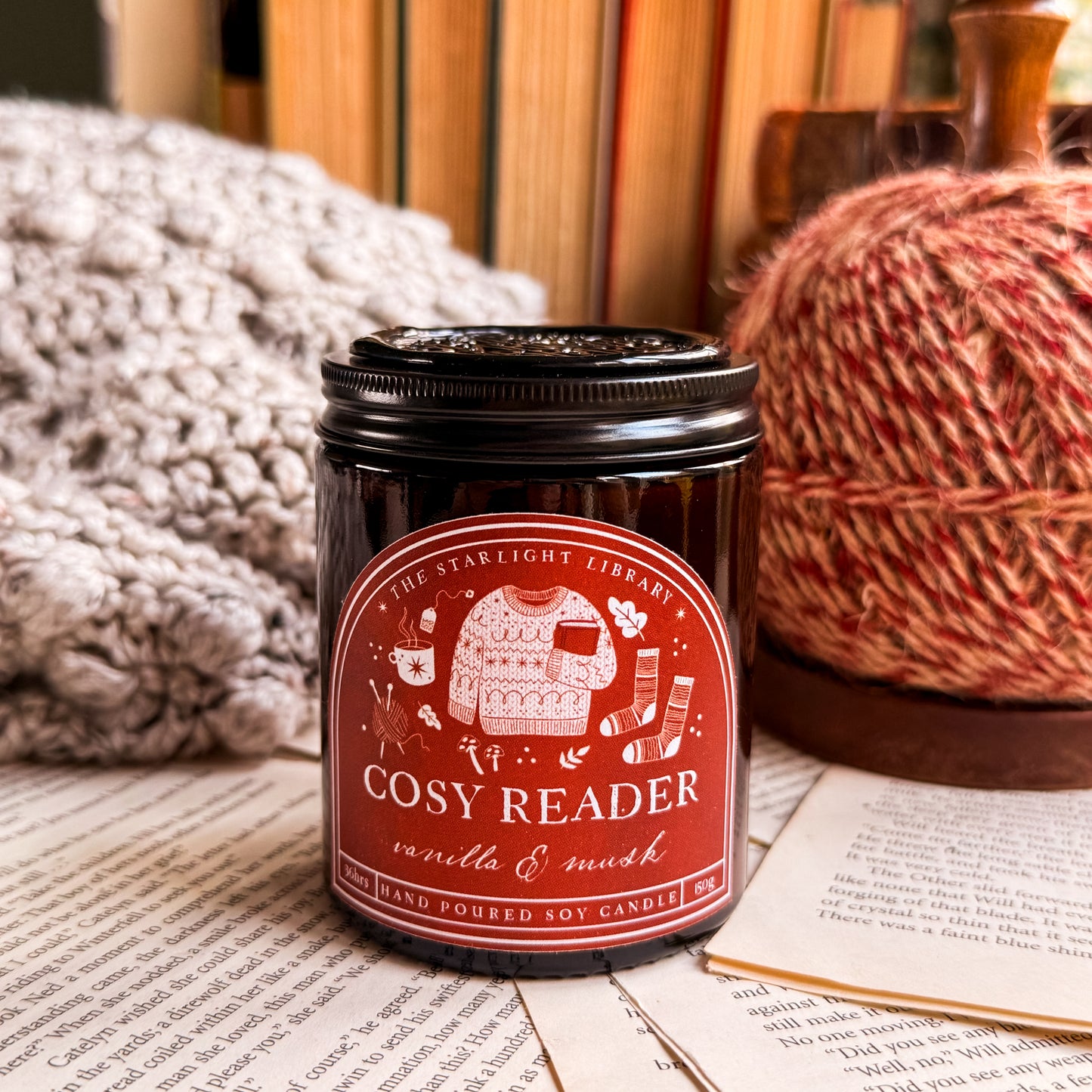 A Cosy Reader candle, with a black lid on and surrounded by pages and cosy knits.