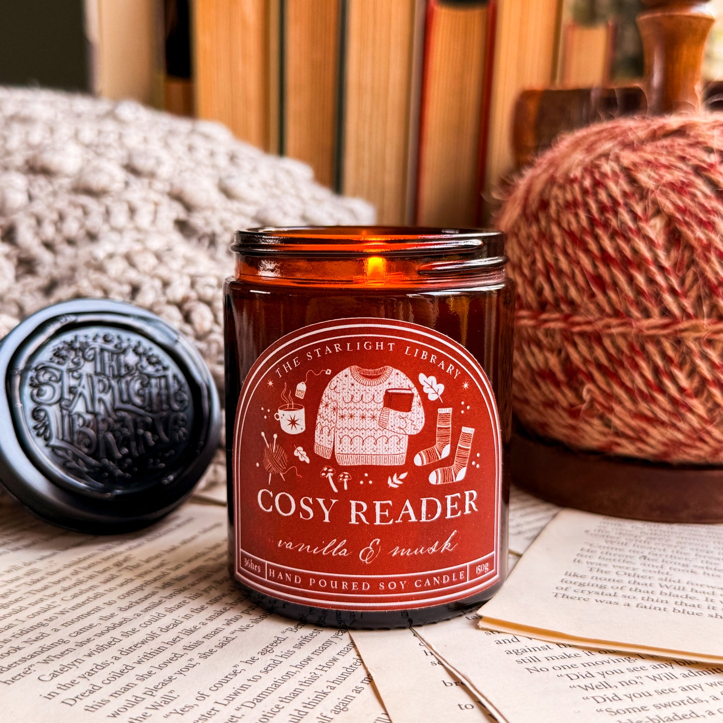 A Cosy Reader candle, lit and surrounded by pages and cosy knits.