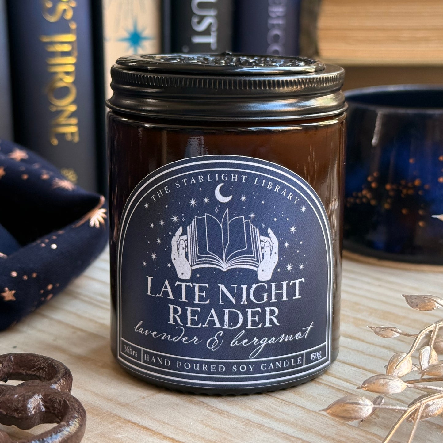 A Late Night Reader candle with a matte black aluminium lid on, topped with a black wax seal. It's surrounded by books and starry accessories.