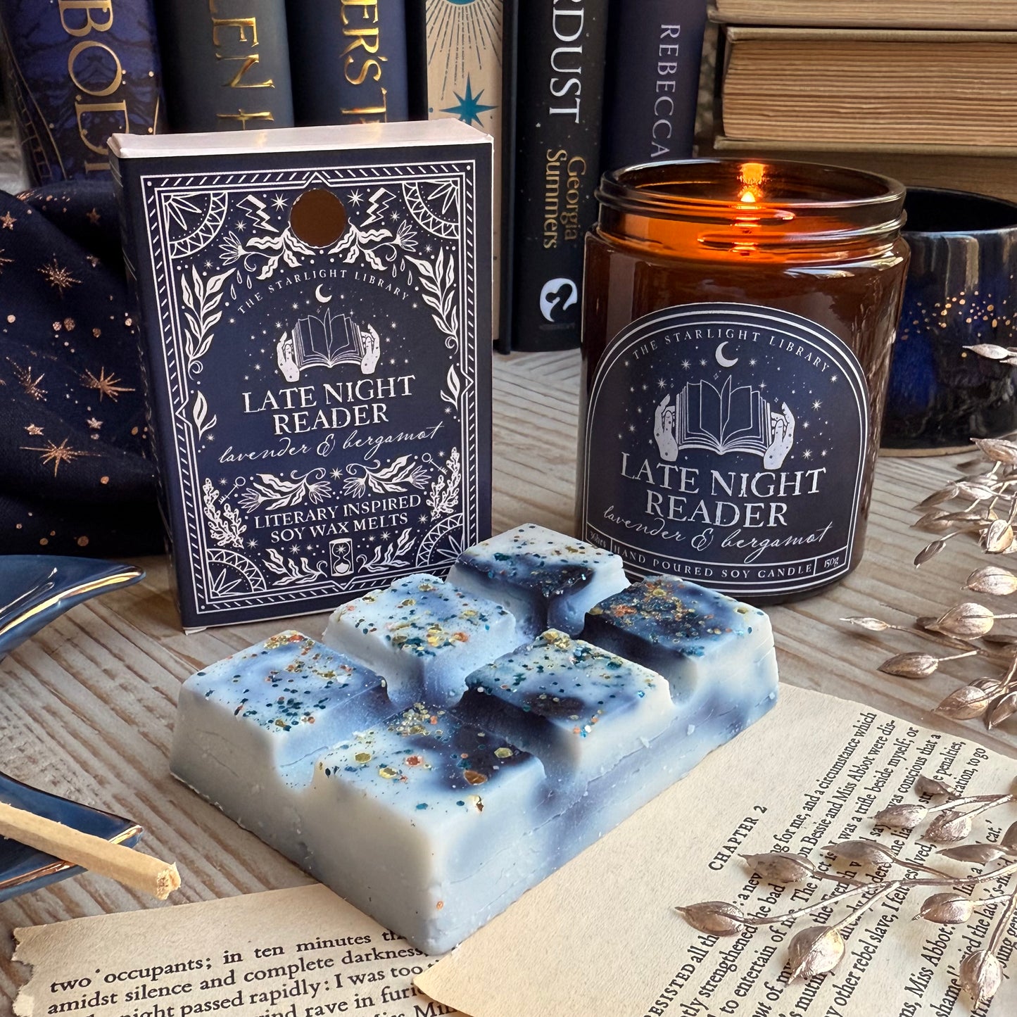 A Late Night Reader wax melt book box, next to a lit Late Night Reader candle. The wax melt bar is unpacked and laid in front. It has inky blue swirls and topped in metallic glitter. They are surrounded by books, pages and starry accessories.
