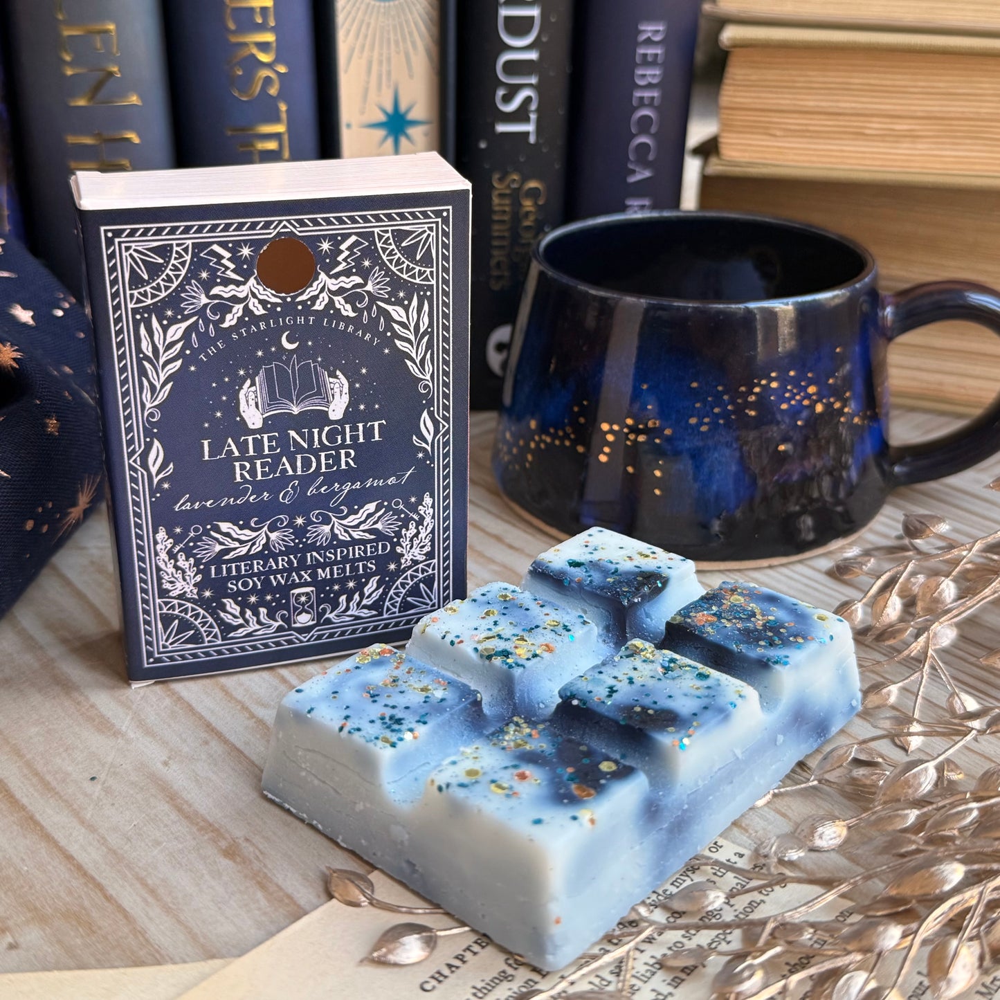 A Late Night Reader wax melt book box, next to a celestial coffee mug. The wax melt bar is unpacked and laid in front. It has inky blue swirls and is topped with metallic glitter. They are surrounded by books and pages and starry accessories.