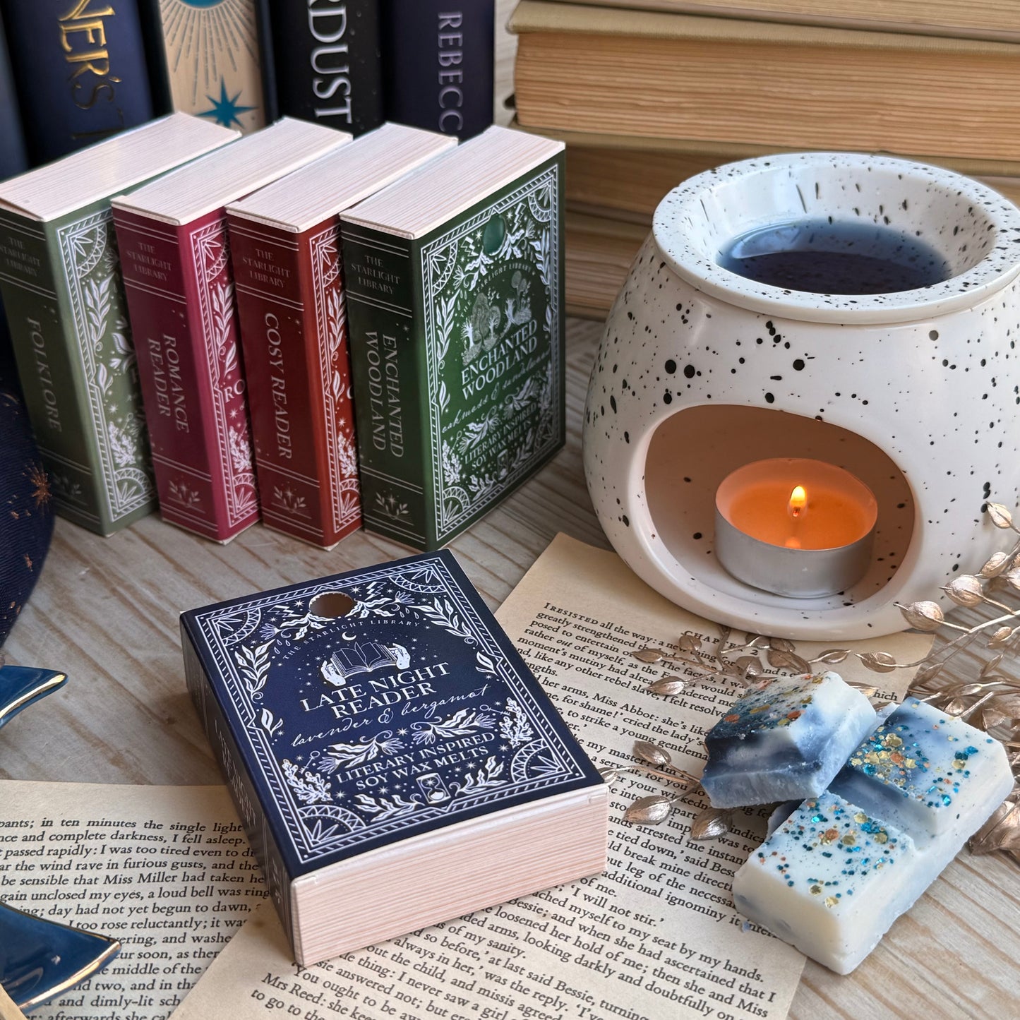 A Late Night Reader wax melt book box, next to a white speckled wax melter. The wax melt bar is unpacked, broekn squares of it laid in front. It has inky blue swirls and is topped with metallic glitter. Behind is a row of other wax melt book boxes, in different colours for different fragrances. They are surrounded by books and pages and starry accessories.