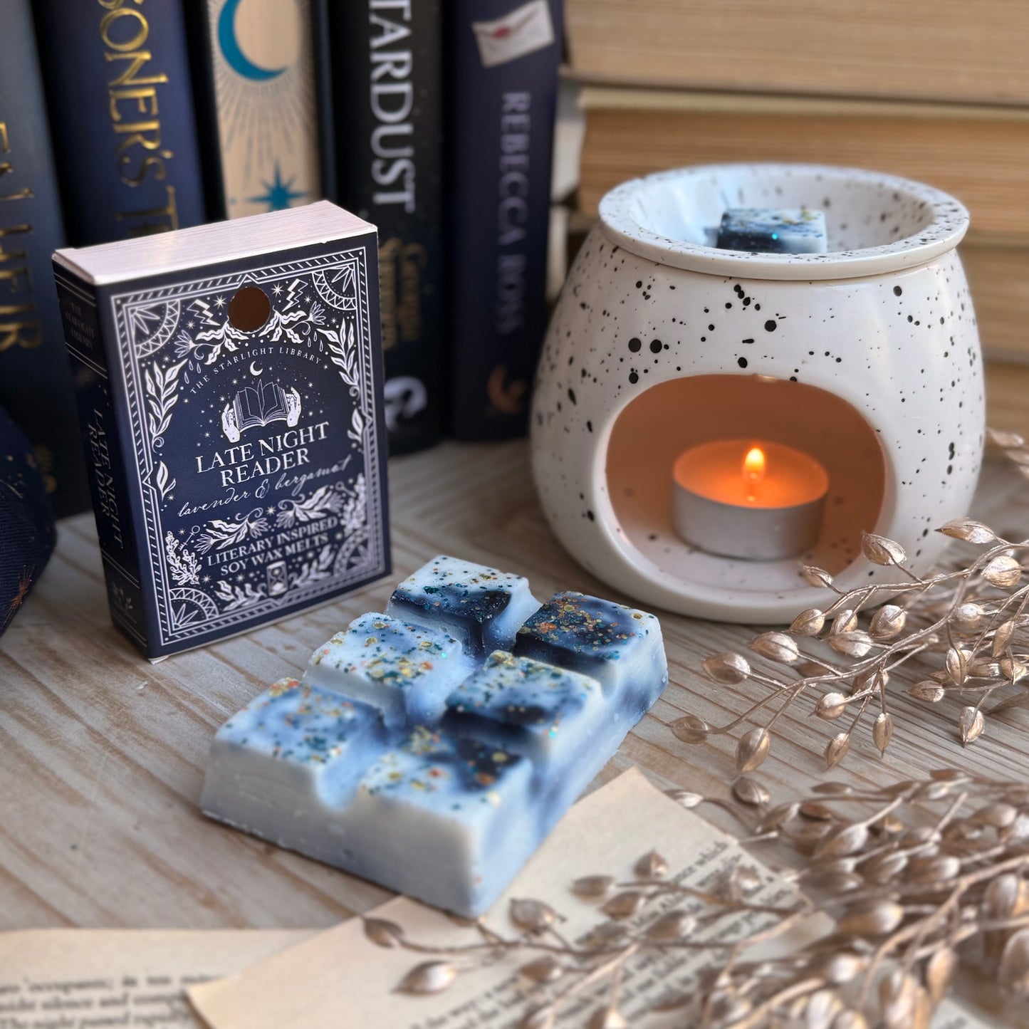 A Late Night Reader wax melt book box, next to a white speckled wax melter. The wax melt bar is unpacked and laid in front. It has inky blue swirls and is topped with metallic glitter. They are surrounded by books and pages and starry accessories.