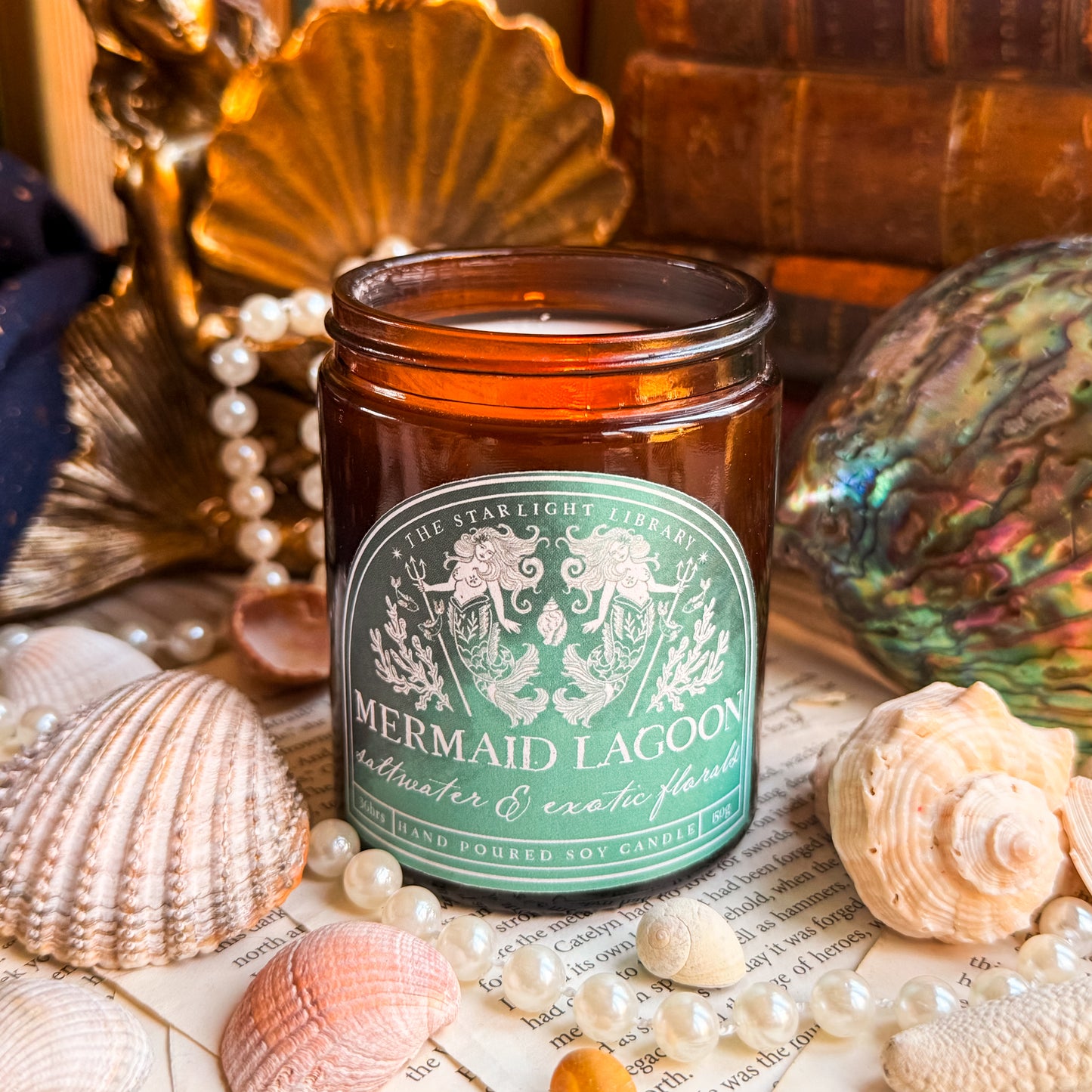 A Mermaid Lagoon candle surrounded by old books, shells and pearls.
