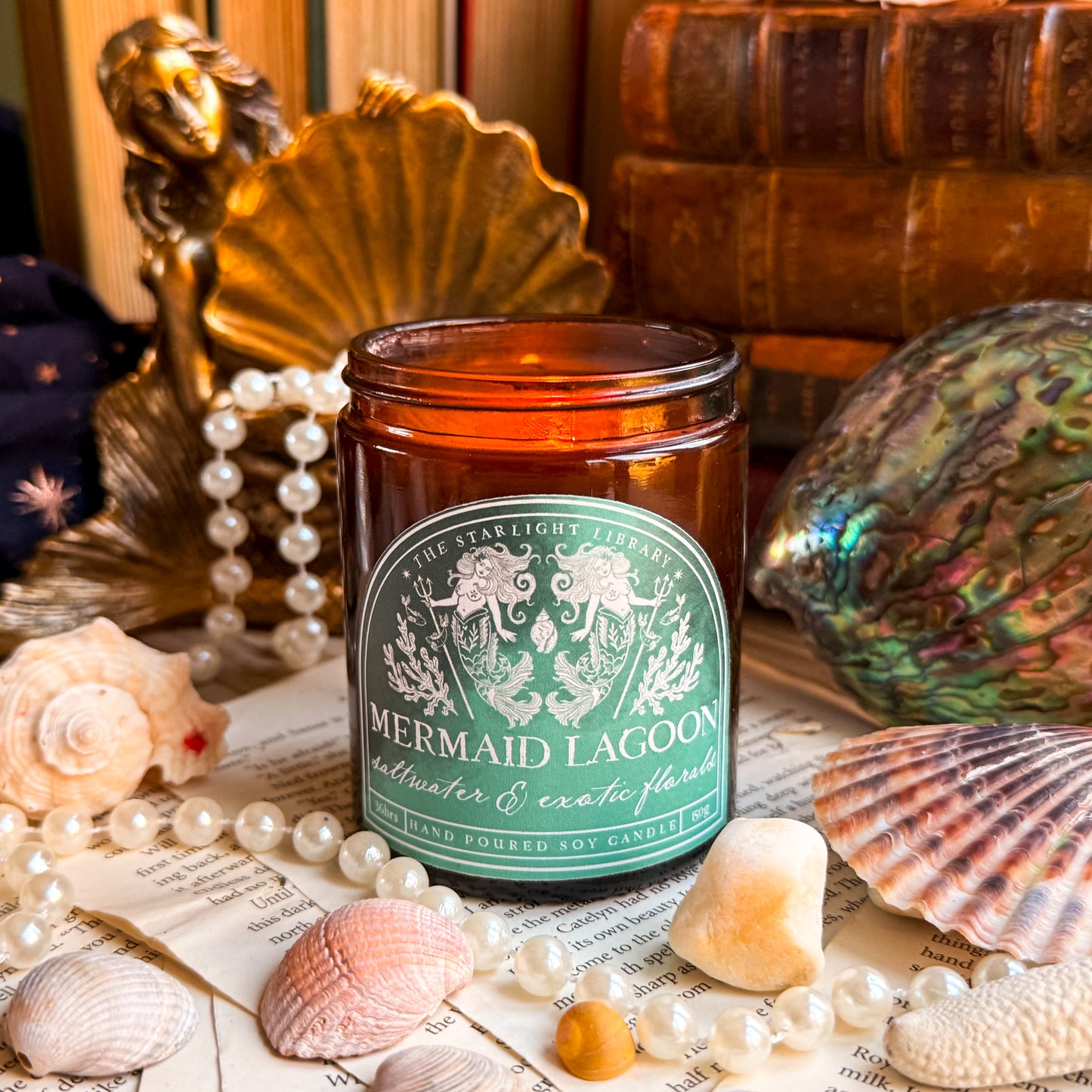 A Mermaid Lagoon candle surrounded by old books, shells and pearls.