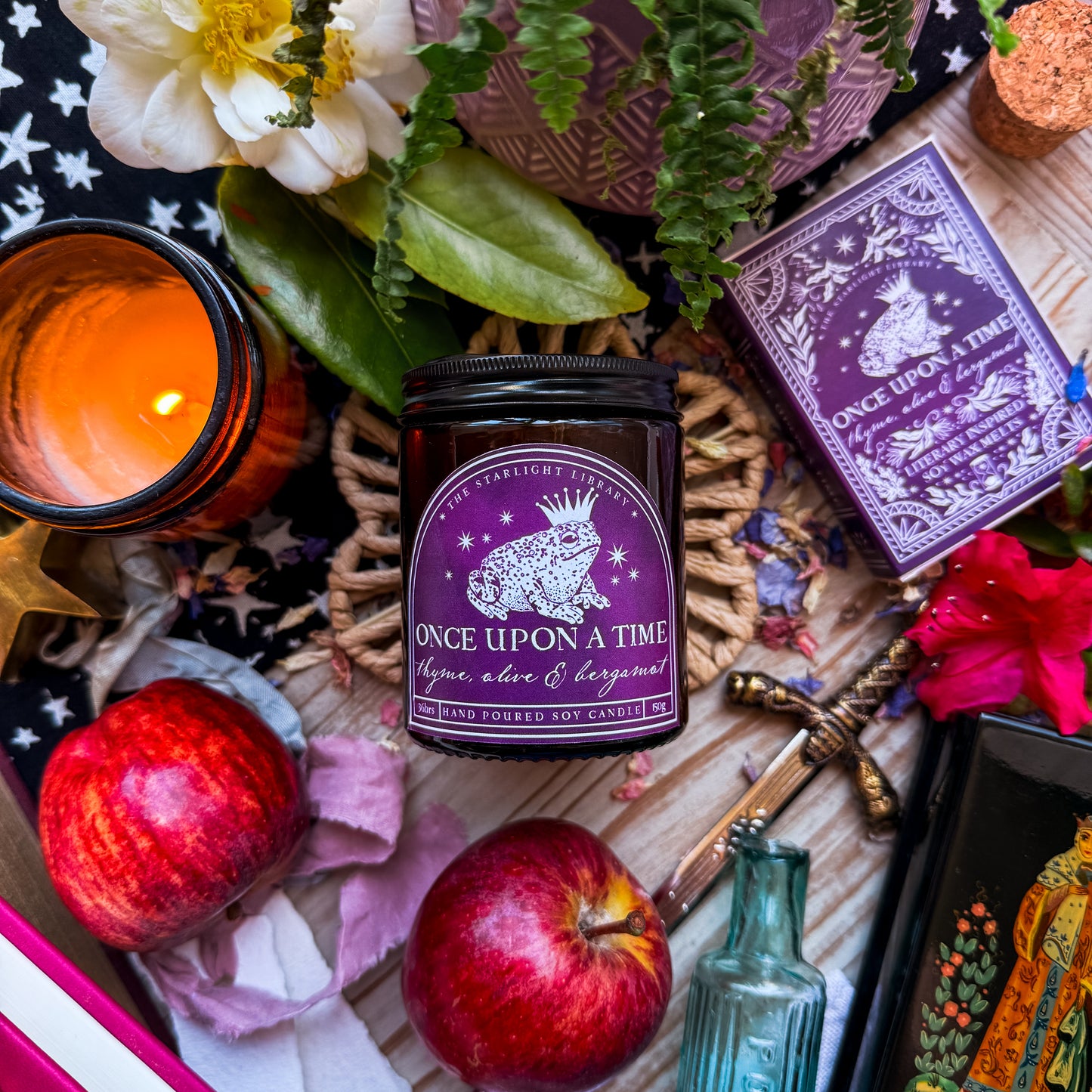 A flatlay of a Once Upon A Time Literary Candle. The candle is surrounded by florals, apples and fairytale iconography.