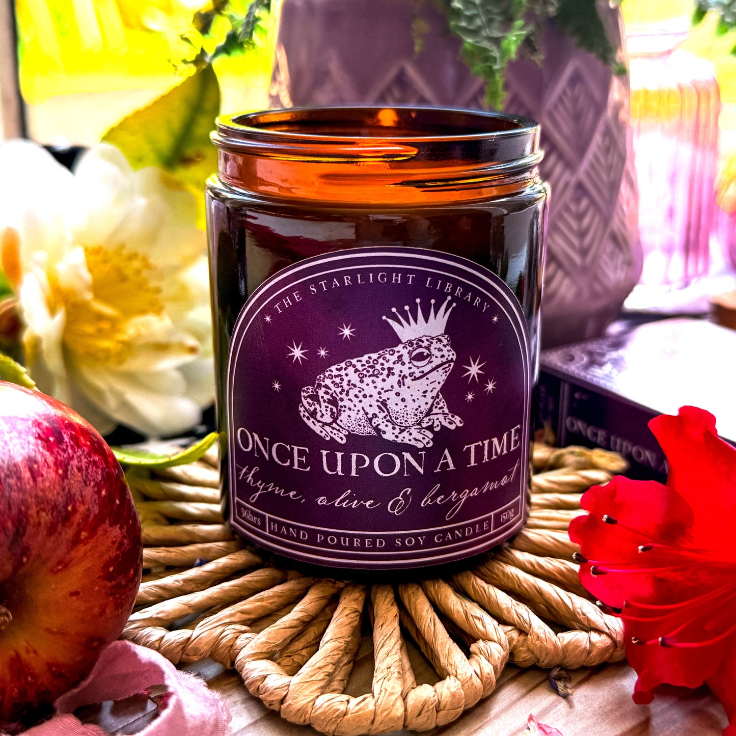 Once Upon A Time Literary Candle. The candle is lit, features a purple label with a toad prince and is surrounded by florals.