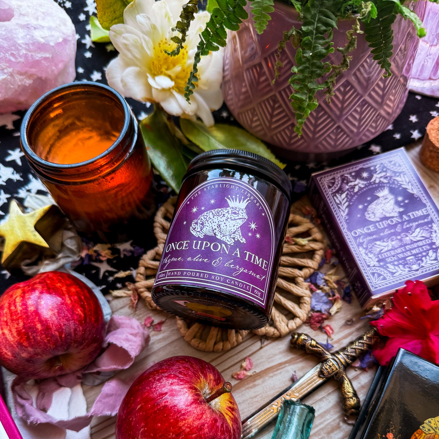 A flatlay of a Once Upon A Time Literary Candle. The candle is surrounded by florals, apples and fairytale iconography.