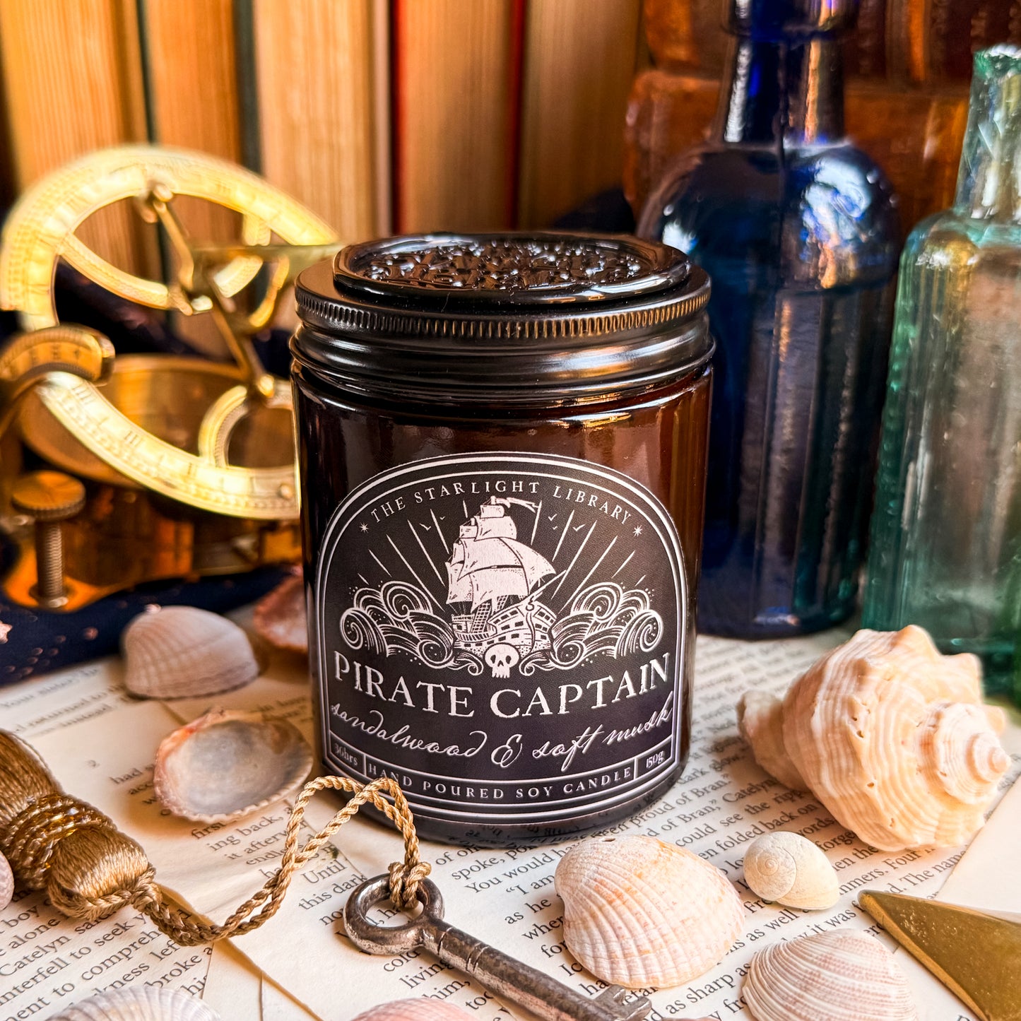 A Pirate Captain candle, surrounded by shells, pages and glass bottles. It has a black lid on with a black wax seal atop.
