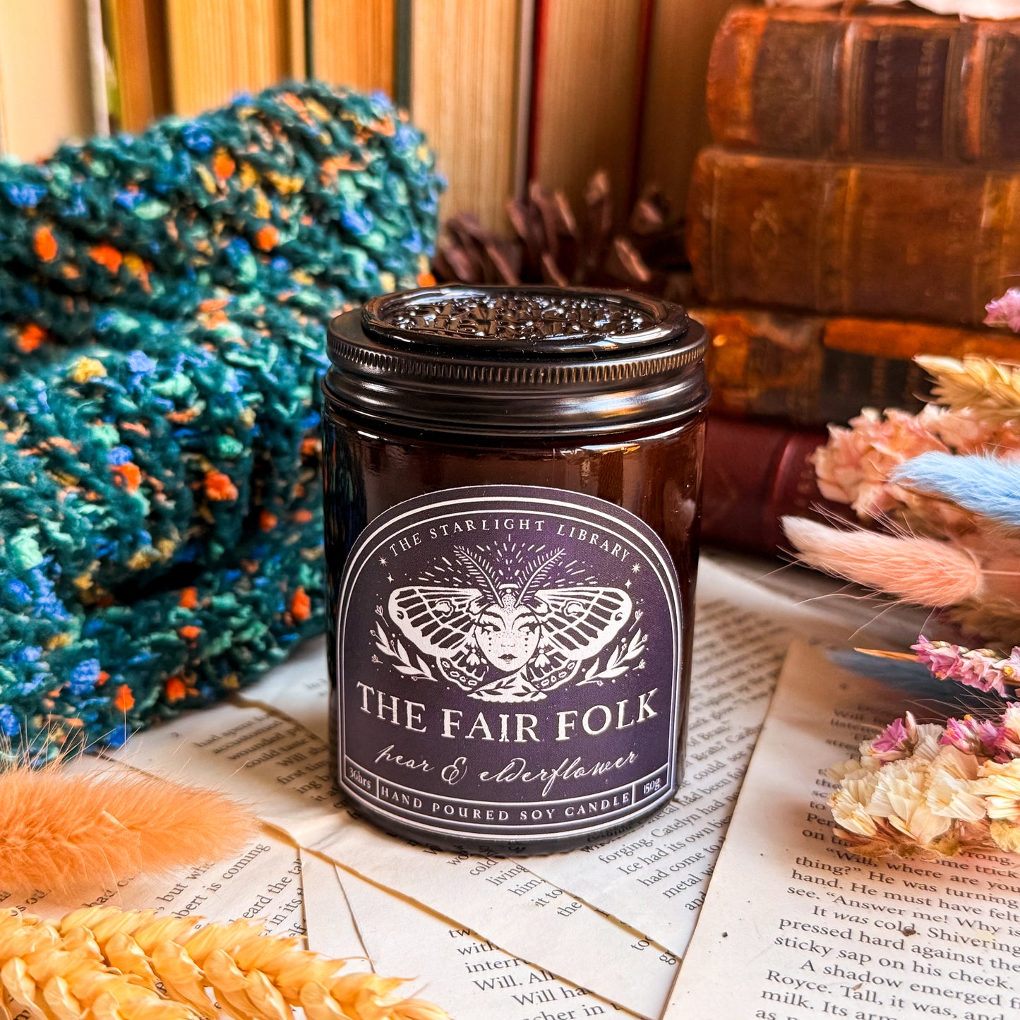 A Fair Folk candle, surrounded by pages, old books and dried flowers. It's topped with a black lid with a black wax seal.