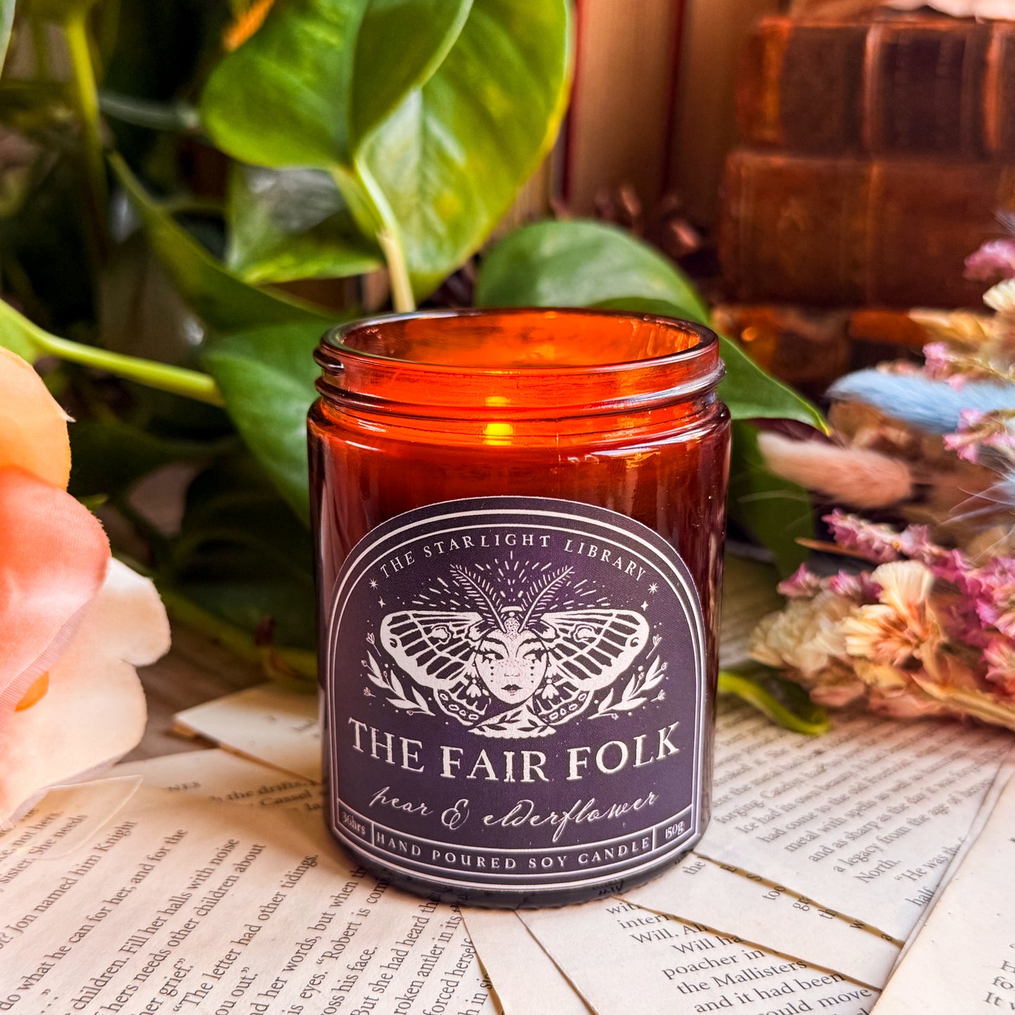 A Fair Folk candle, lit, surrounded by pages, old books and dried flowers.