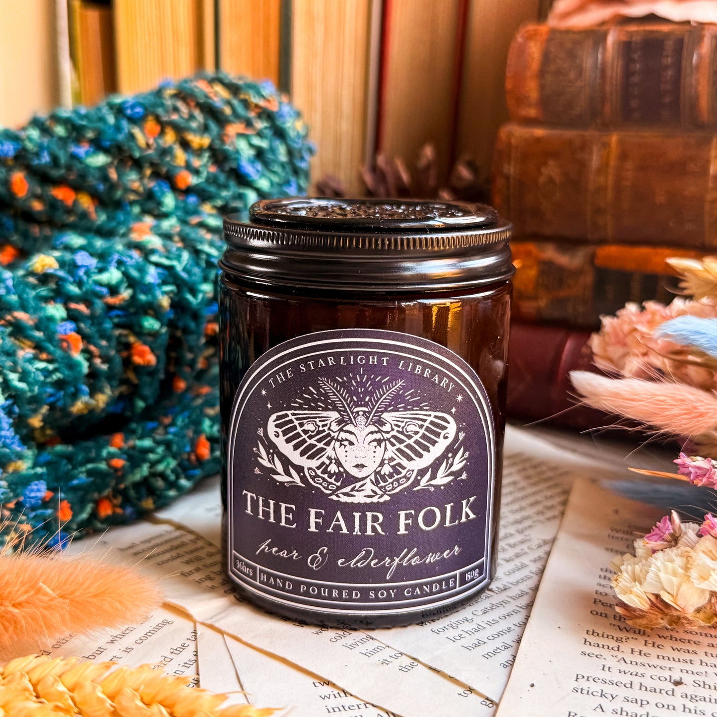 A Fair Folk candle, surrounded by pages, old books and dried flowers. It's topped with a black lid with a black wax seal.