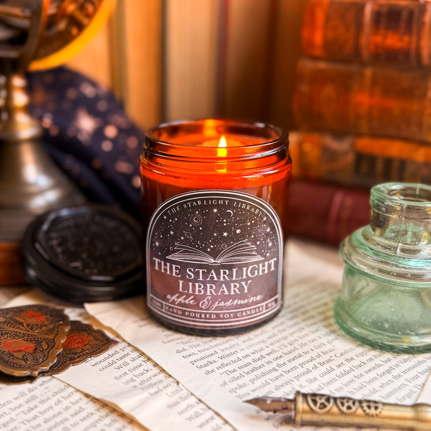 The Starlight Library signature candle.The candle is lit and surrounded by books and book pages.