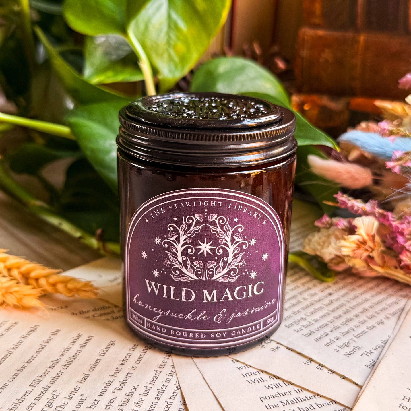 A Wild Magic candle, with a black lid topped with a black wax seal. It is surrounded by book pages, leaves and dried flowers.
