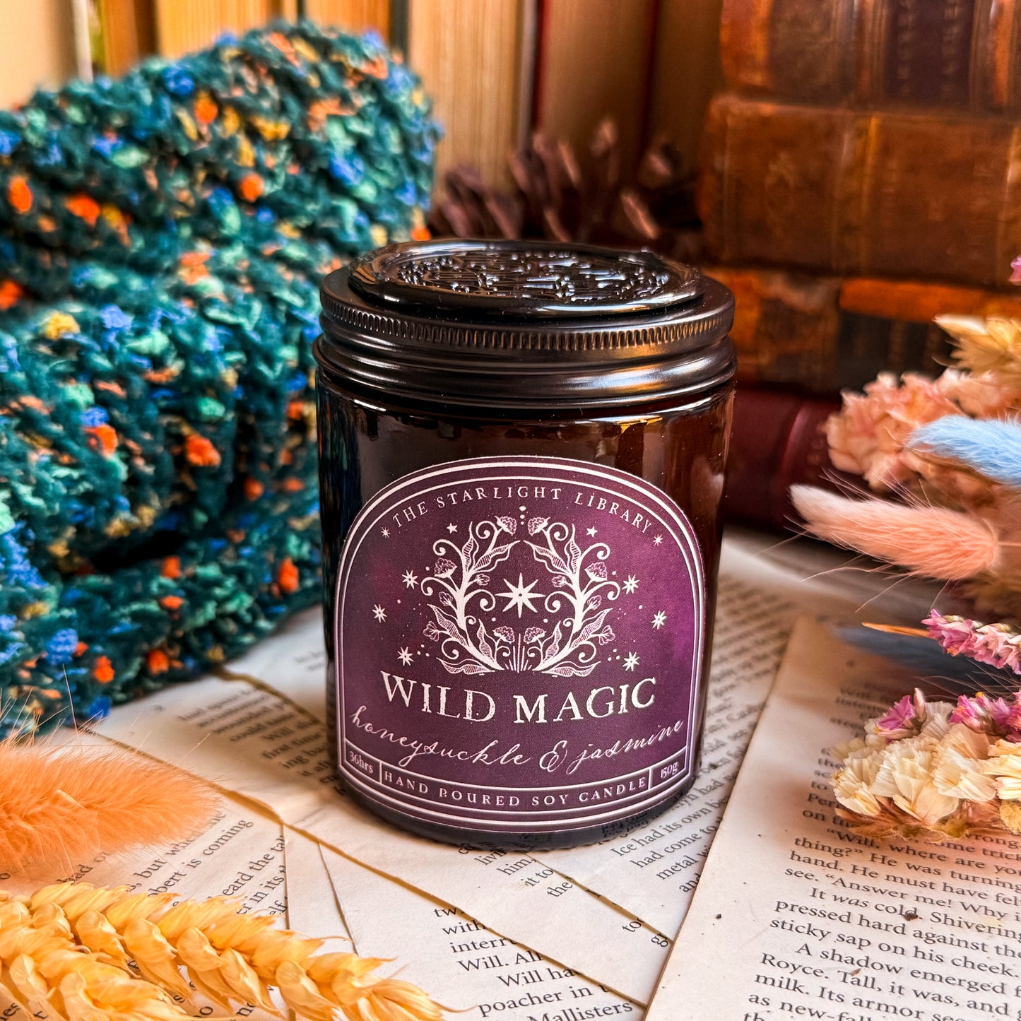 A Wild Magic candle, with a black lid topped with a black wax seal. It is surrounded by book pages, cosy knits and dried flowers.