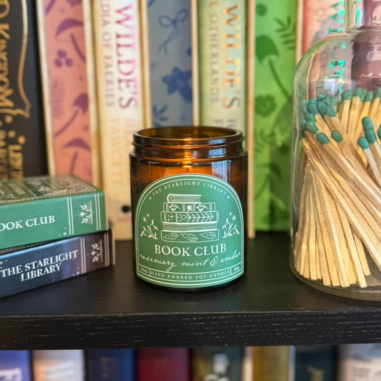 A Book Club candle, on a bookshelf. It has a background of colourful sprayed edges and book spines, and a pot of long green matches next to it.