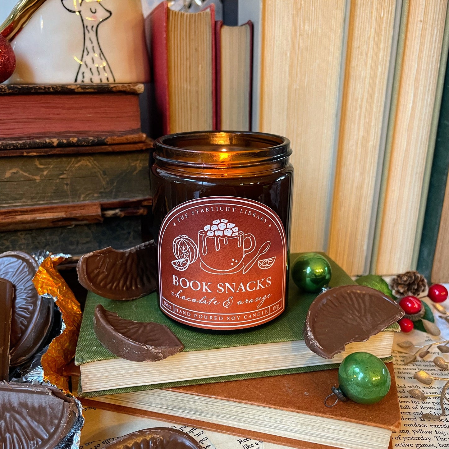Candle labeled 'Book Snacks' on a stack of books with chocolate snacks and decorative items.