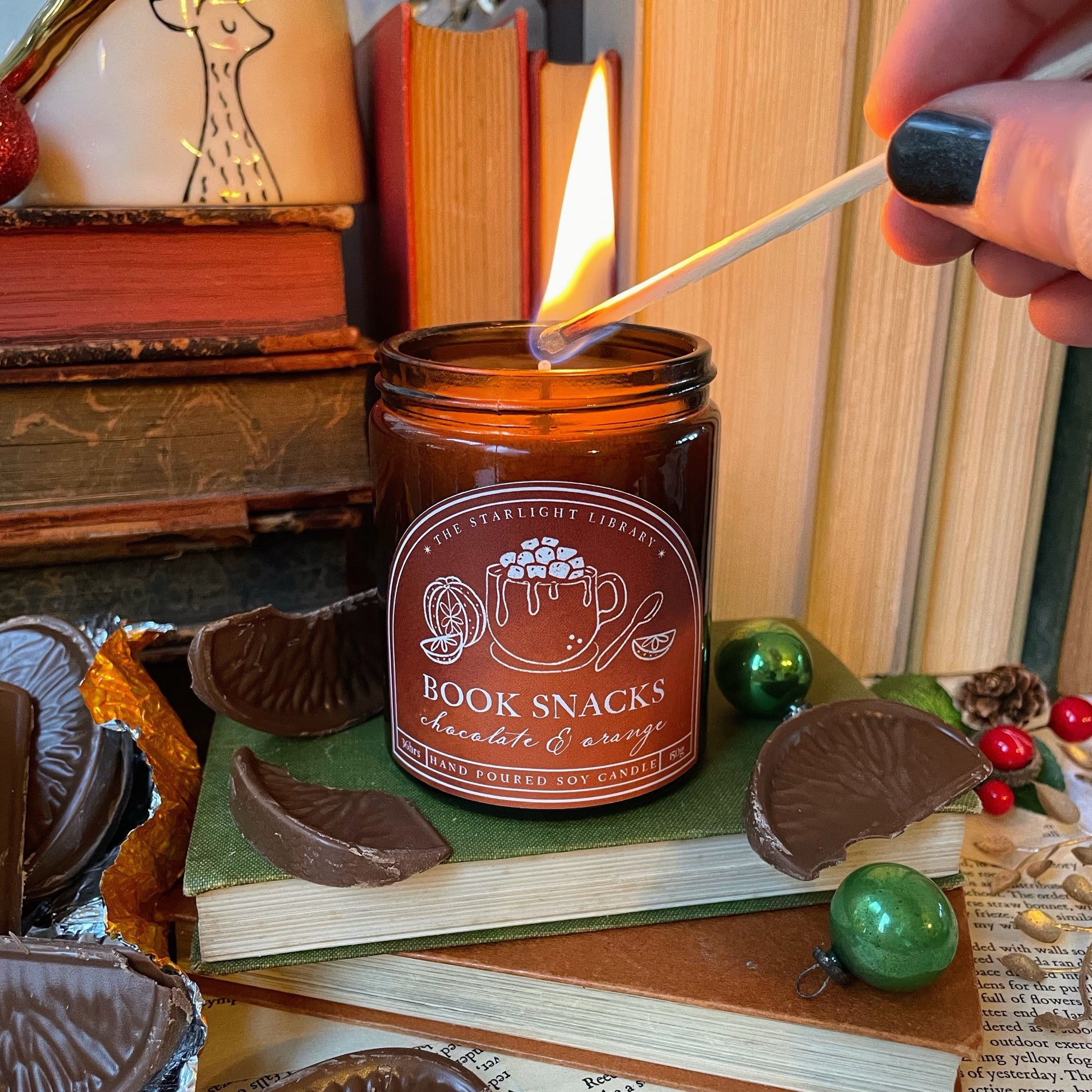 Candle labeled 'Book Snacks' being lit with a match, surrounded by chocolate snacks and books.