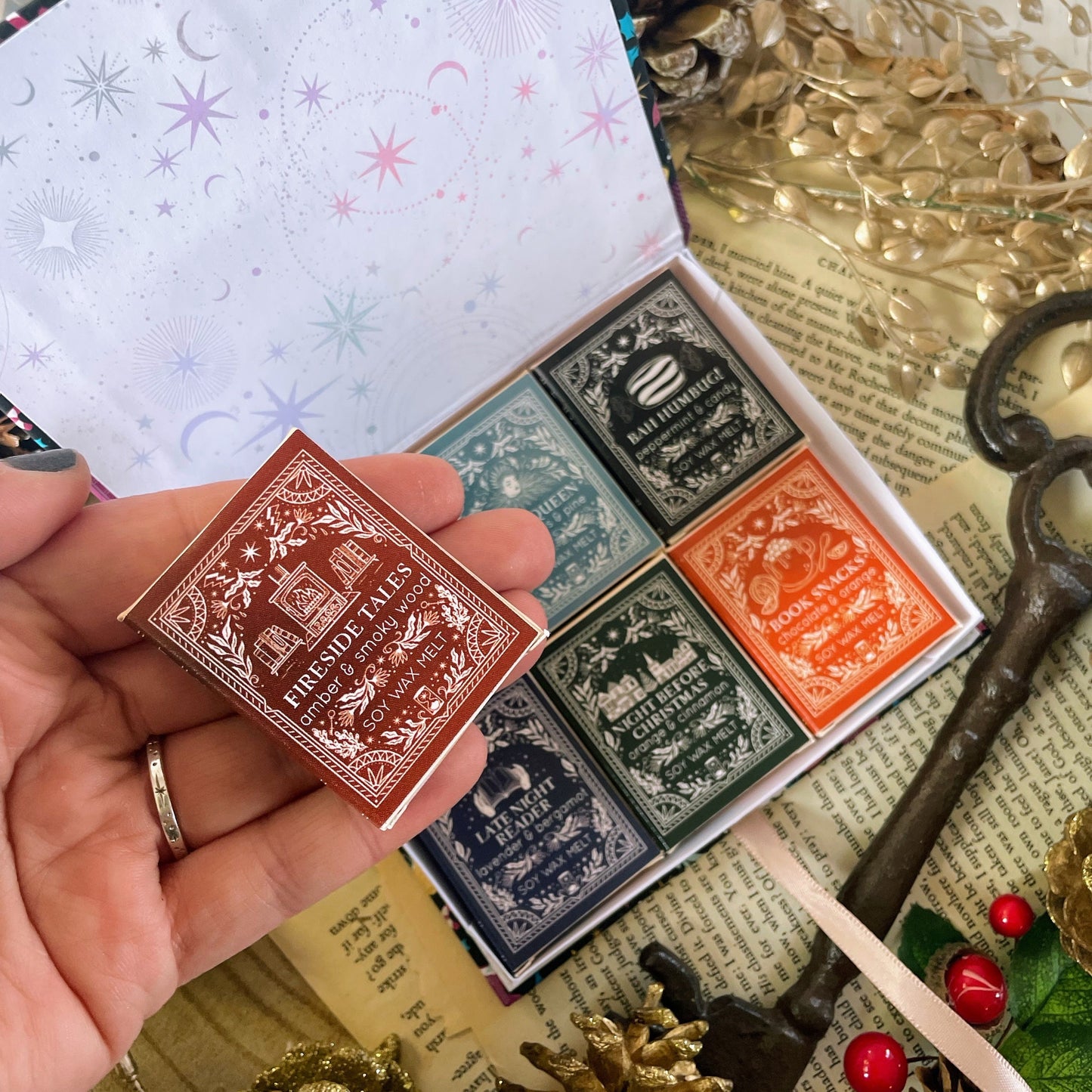Collection of mini wax melt book boxes with decorative labels held by a hand, on a textured surface with decorative elements.