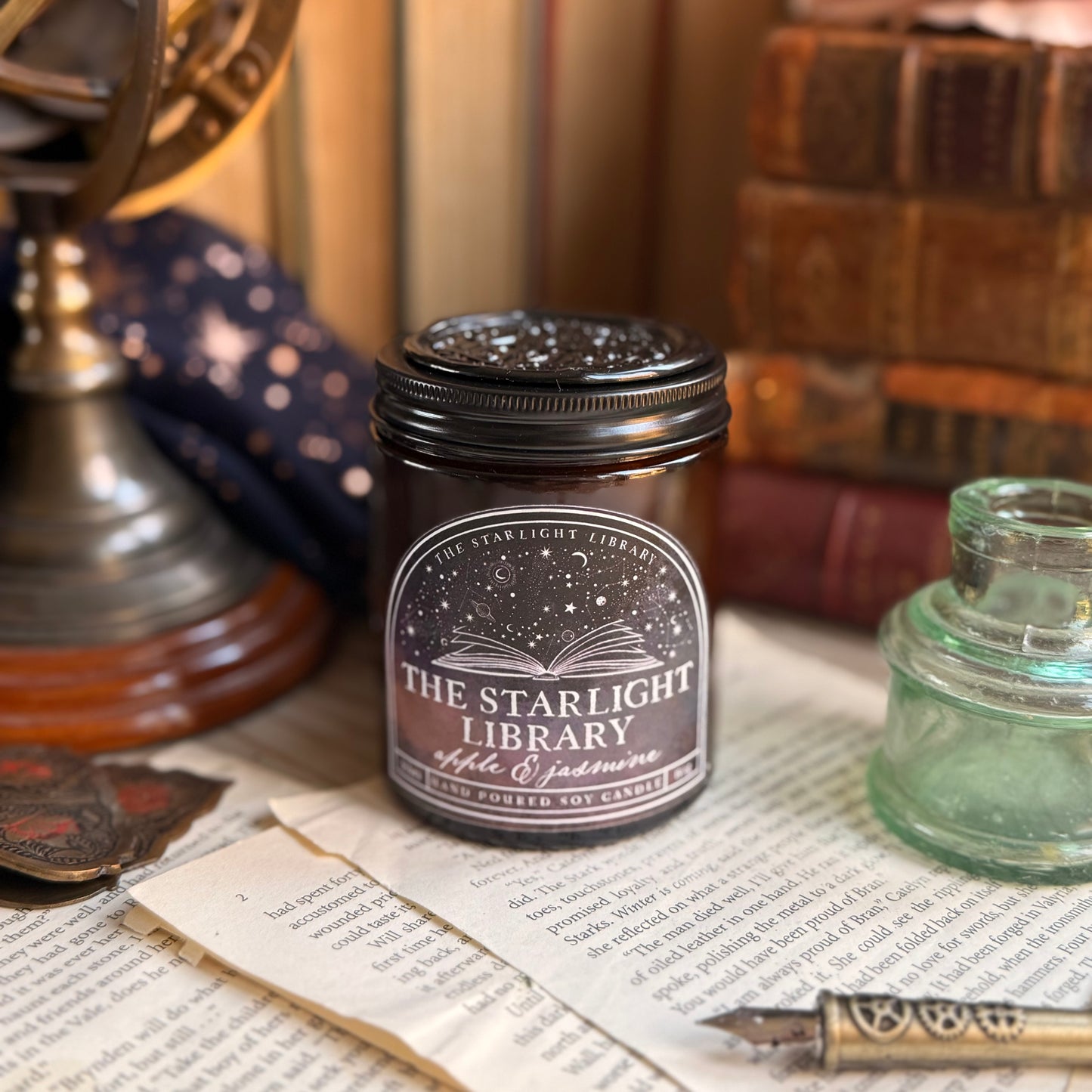 The Starlight Library signature candle.The candle has a black aluminium lid with a black wax seal on top and is surrounded by books and book pages.