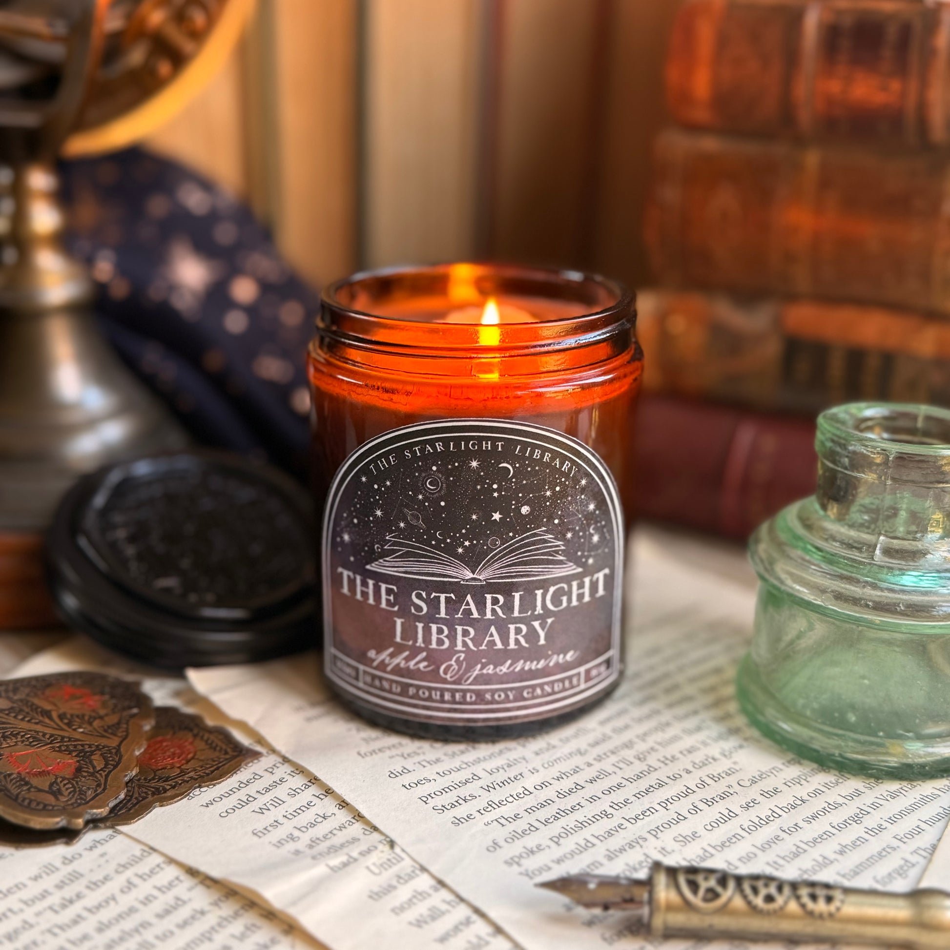The Starlight Library signature candle.The candle is lit and surrounded by books and book pages.