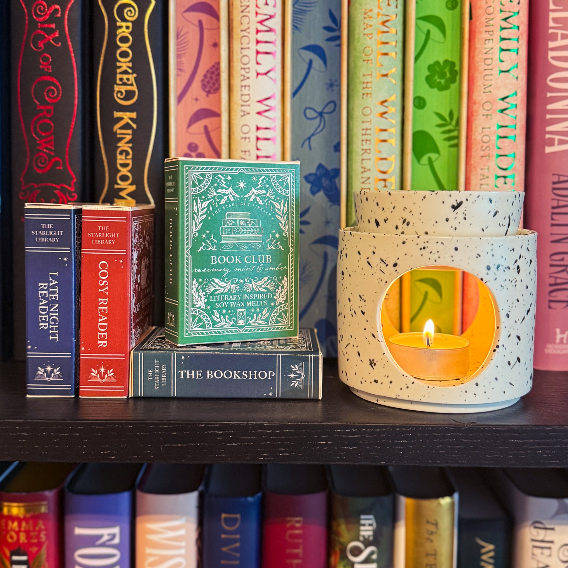 Wax Melt burner with a lit candle beneath, on a bookshelf with various books and a stack of wax melt book bars including Book Club.